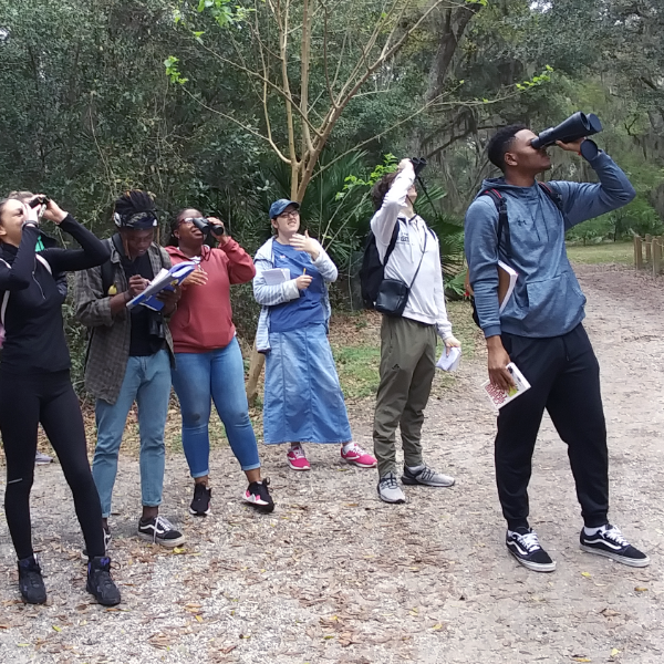 A group of six student outdoors near trees looking up at the sky through binoculars.